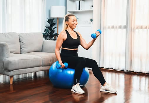 Athletic and sporty senior woman sitting on fit ball while engaging in weight lifting with dumbbell at home exercise as concept of healthy fit body lifestyle after retirement. Clout