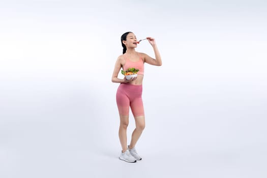 Young sporty Asian woman in sportswear holding salad bowl fill with vibrant of fruit and vegetable. Natural youthful and fit body lifestyle with balance nutrition on isolated background. Vigorous