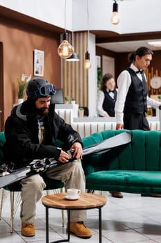 Male tourist in winter gear sits on cozy couch in hotel lobby, securing his wintersports gear. Caucasian man maintaining his skiing skis, preparing for a thrilling snowy slopes mountain experience.