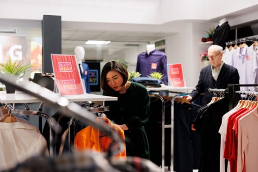 People customers choosing clothes while doing purchases in fashion mall during seasonal sales. Asian woman shopper looking through clothing on hangers while shopping on Black Friday