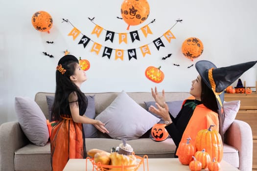 A playful young mother sits on a comfortable sofa in the living room decorated with pumpkins and her cute little daughter tells a spooky Halloween story. Funny mother and girl having fun at home.