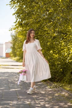 Beautiful girl with flowers, in a dress, glasses and sneakers. A beautiful bouquet of flowers in a box in the hands of a beautiful girl who walks along the street on a sunny day.