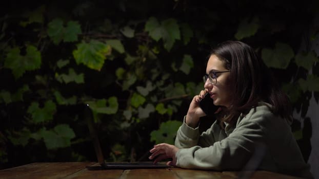 A young woman at a table in a gazebo talking on the phone late at night. A girl in a hoodie and glasses with a phone and laptop on a background of green grapes. 4k