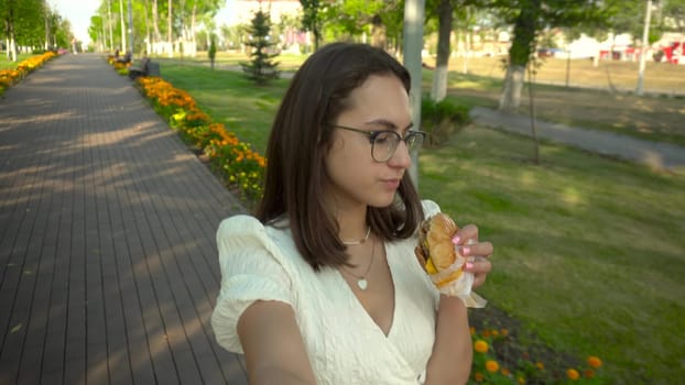 A young woman walks along an alley in a white dress and eats a burger. A girl with glasses walks through the park and films herself. 4k