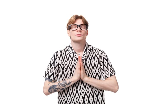 young european male student with red hair dressed in a shirt with a diamond pattern shows experiencing calmness.