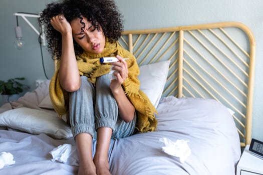 African American young woman checking temperature with thermometer, sitting in bed feeling unwell. Woman sick in bed. Sickness concept.