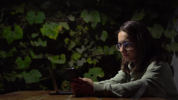 A young woman at a table in a gazebo is texting on a smartphone late at night. A girl in a hoodie and glasses with a phone in her hands against a background of green grapes. 4k