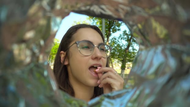 A young woman with glasses eats potato chips from a pack while walking in the park. View of a woman from a bag of chips. 4k