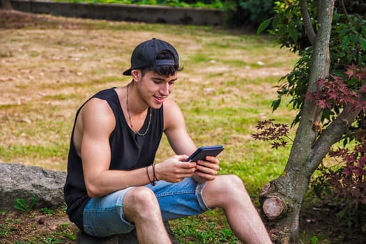 A man sitting on a rock in the grass, reading from an ebook reader. Photo of a handsome young man sitting on a rock in the grass with an e-book reader