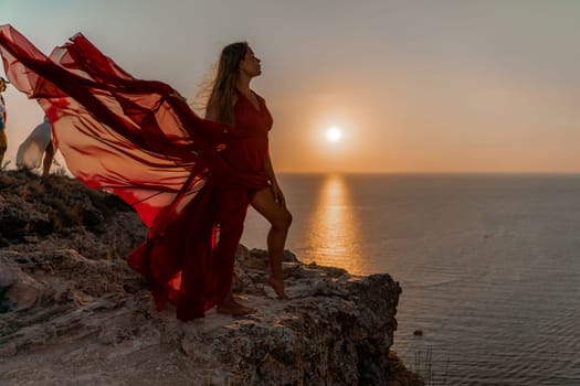 Woman sunset sea red dress, side view a happy beautiful sensual woman in a red long dress posing on a rock high above the sea on sunset