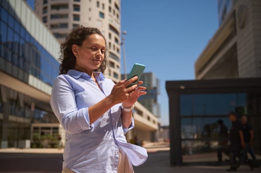 Bottom view of a female entrepreneur holding her modern smartphone, checking new mobile app, booking, ordering, browsing web sites, making online reservations, standing outdoors