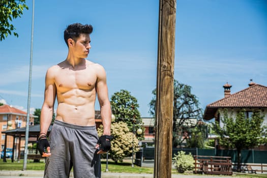 A man without a shirt standing next to a pole, in a summer day, ready to workout outdoor