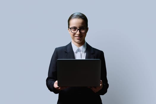 Teenage female student in formal business style using computer laptop, on grey studio background. Digital technologies, education, virtual educational services, online learning, youth concept