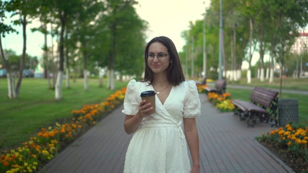 A young woman walks along an alley in a white dress and drinks crofe. A girl with glasses walks through the park with coffee in her hands. 4k