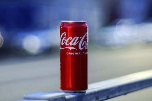 Tyumen, Russia-August 10, 2024: can of soft drink coca cola sits on a railing as city lights blur in the background during early evening