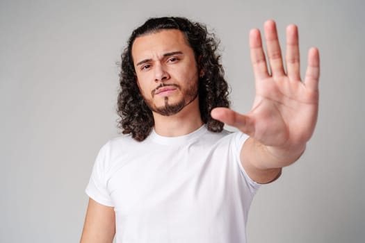 Young man with curly hair gestures to stop while wearing a plain white t-shirt against a neutral background close up