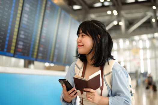 Young woman holding a passport and using mobile phone for checking flight information in an airport.
