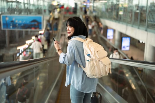 Happy young woman with backpack using mobile phone while on escalator in an airport terminal.