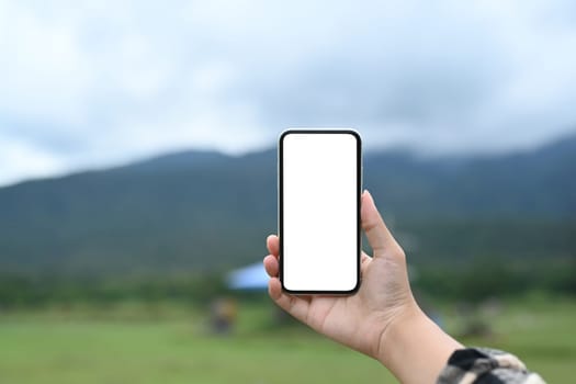 Hand holding smartphone with white screen on blurred background of mountains landscape.