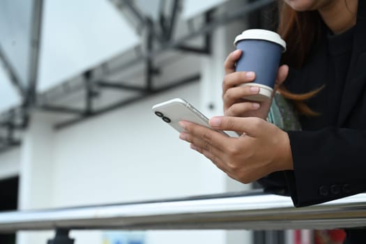 Young businesswoman with a cup of coffee leaning on the railing and using mobile phone.