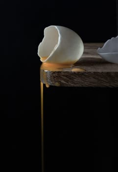 Conceptual image of a broken white eggshell with dripping egg yolk on black background. A cracked raw white duck egg shell and dripping yolk on the edge of a wooden cutting board and dark background, use it as your Wallpaper, Poster and Copy space, Selective focus.