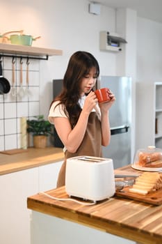 Pretty young woman drinking coffee while preparing the breakfast in the kitchen.