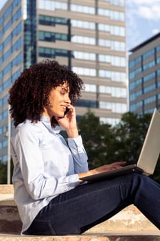 side view of focused happy female entrepreneur sitting on stone bench in park and working remotely while speaking on smartphone and using laptop