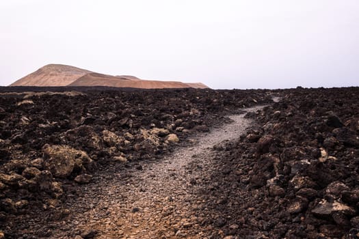 rough route with trail between dark lava stones against Caldera Blanca and Caldereta volcano in Natural Park of Lanzarote in Canary Islands, Spain, concept of volcanic landscape and wild nature