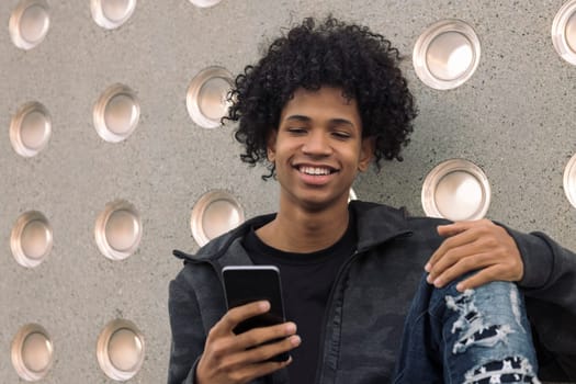 smiling african american hipster male teenager with curly hair using mobile phone while sitting near concrete wall, concept of technology of communication and urban lifestyle