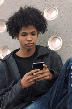 serious african american hipster male teenager with curly hair using mobile phone while sitting near concrete wall, concept of technology of communication and urban lifestyle