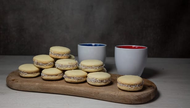 A stack of tasty alfajores cookies with coconut filling displayed on a wooden board, accompanied by two ceramic coffee cups, creating a cozy atmosphere for enjoying a sweet treat.