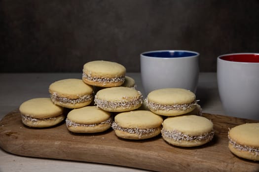 A stack of tasty alfajores cookies with coconut filling displayed on a wooden board, accompanied by two ceramic coffee cups, creating a cozy atmosphere for enjoying a sweet treat.
