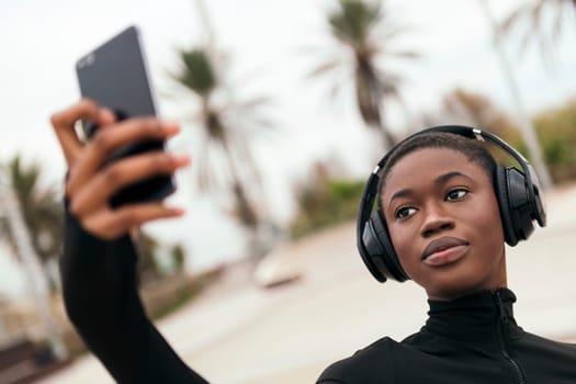 young ethnic female in casual apparel with wireless headset taking self portrait on mobile phone in a park
