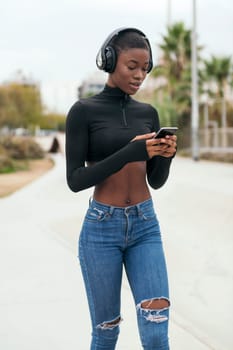 young glad african american female in wireless headphones using mobile phone while listening to music in city