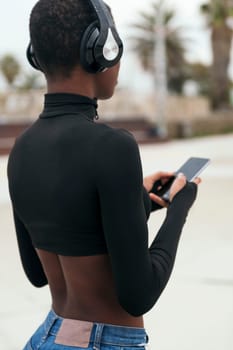 back view of african american female with headphones text messaging on mobile phone with black screen in city