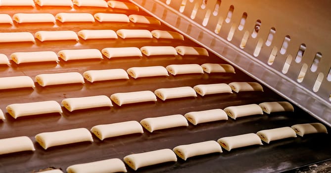 Rows of uncooked bread rolls are neatly aligned on a factory conveyor belt, ready for baking. The machinery suggests a large-scale production in an industrial bakery setting, emphasizing efficiency in food manufacturing. Golden dough shapes are progressing through the initial phases of a baking process.