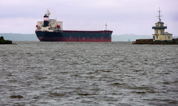 In a busy harbor, a large cargo vessel sails through tranquil waters, accompanied by an iconic lighthouse under a cloudy sky. The scene captures maritime activity.