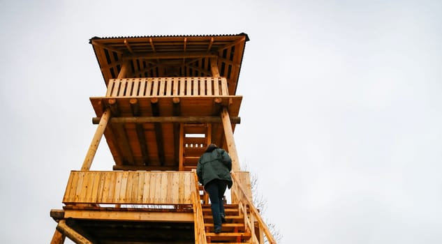 A person ascends the wooden stairs of a lookout tower in a rural landscape. The cloudy sky provides a serene backdrop for the climb.
