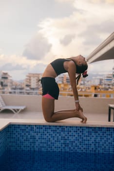 A woman elegantly performs a beautiful yoga pose by a pristine and serene swimming pool, framed by a dramatic skyline