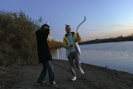 Two friends stroll along the water edge at twilight, wearing festive hats and sharing laughter.