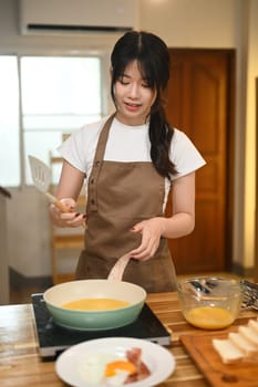 Young woman wearing a brown apron cooking scrambled eggs in a green frying pan.