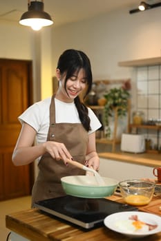 Cheerful young asian woman cooking scrambled eggs in frying pan on a stovetop.