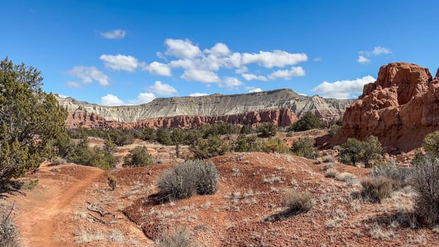 A scenic view of a hiking trail at Kodachrome Basin State Park in Utah. Red rock formations, dramatic cliffs, and desert shrubs and trees are visible beneath a blue sky with white clouds - USA