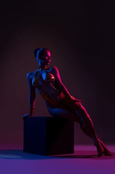 Beautiful nude girl with gold tape body art sitting on a cube in dark room