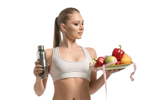 Beautiful fitness trainer holding a plate with friut, a water bottle and a tape-measure cropped isolated view on white background
