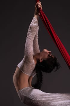 Side view of beautiful charming slim gymnast with upped hands hanging on red material on black background