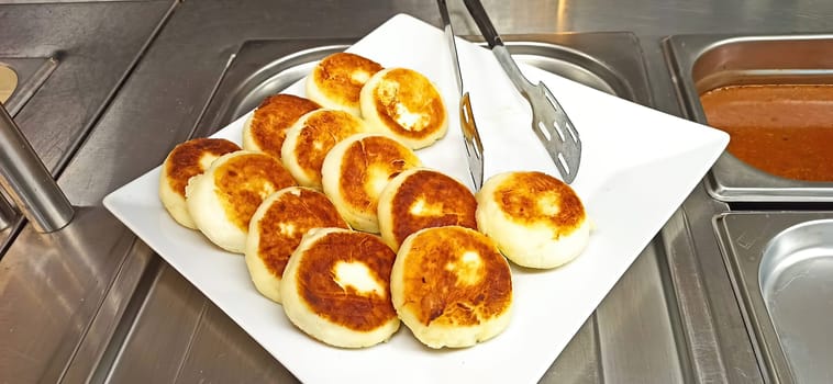 A plate of fried cheese cakes sits on a stainless steel counter. The plate is white and the cheese cakes are golden brown and slightly crispy.