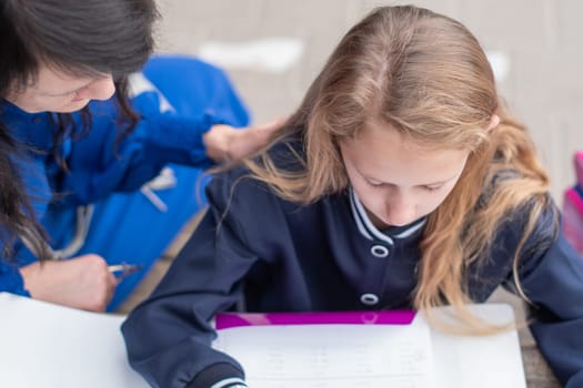 smart girl solving math problems on her own while her mother sits nearby at wooden table on children's playground and watches,education concept,going back to school, girl in beautiful blue uniform