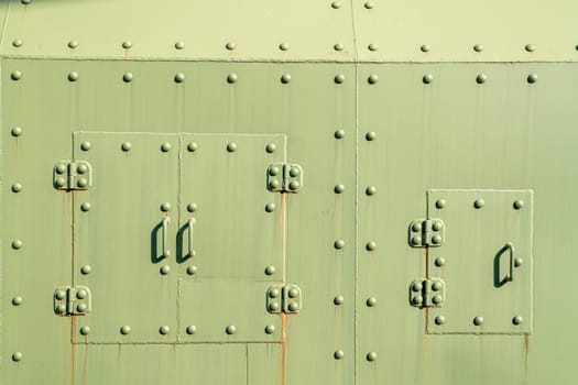 A textured, rusty green metal surface features rivets and two secured panels. The sunlight casts shadows on the surface, highlighting its industrial character and age.