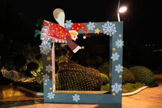 A whimsical Santa Claus figure is positioned above a large decorative frame in a beautifully lit garden. The vibrant colors and festive designs add a joyful atmosphere during the holiday season.